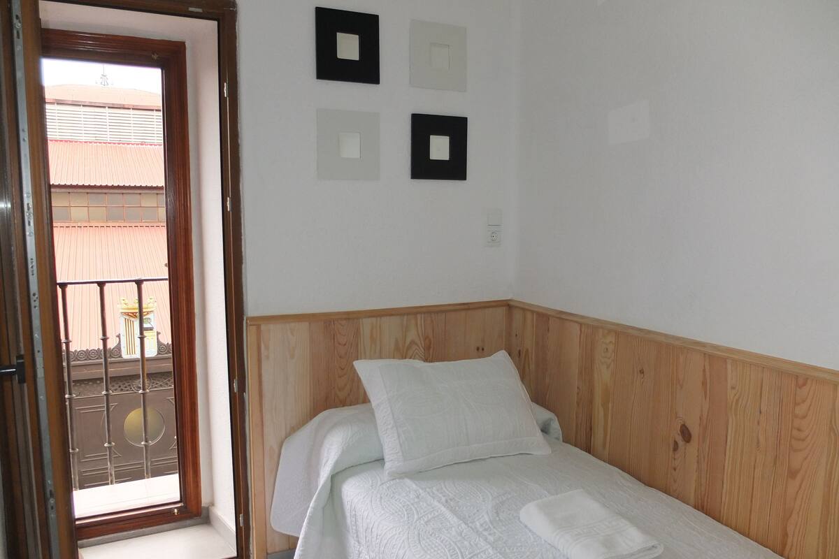 Un lit simple avec une tête de lit en bois, une literie blanche et une petite fenêtre donnant sur un bâtiment avec un toit rouge.