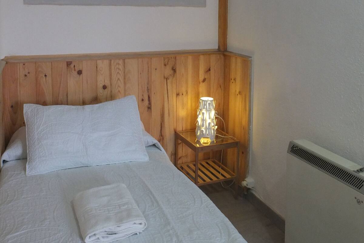 Une chambre d’hôtel avec une tête de lit en bois, un lit recouvert de draps blancs, un tableau représentant un sentier forestier, une table de chevet avec une lampe et un radiateur.