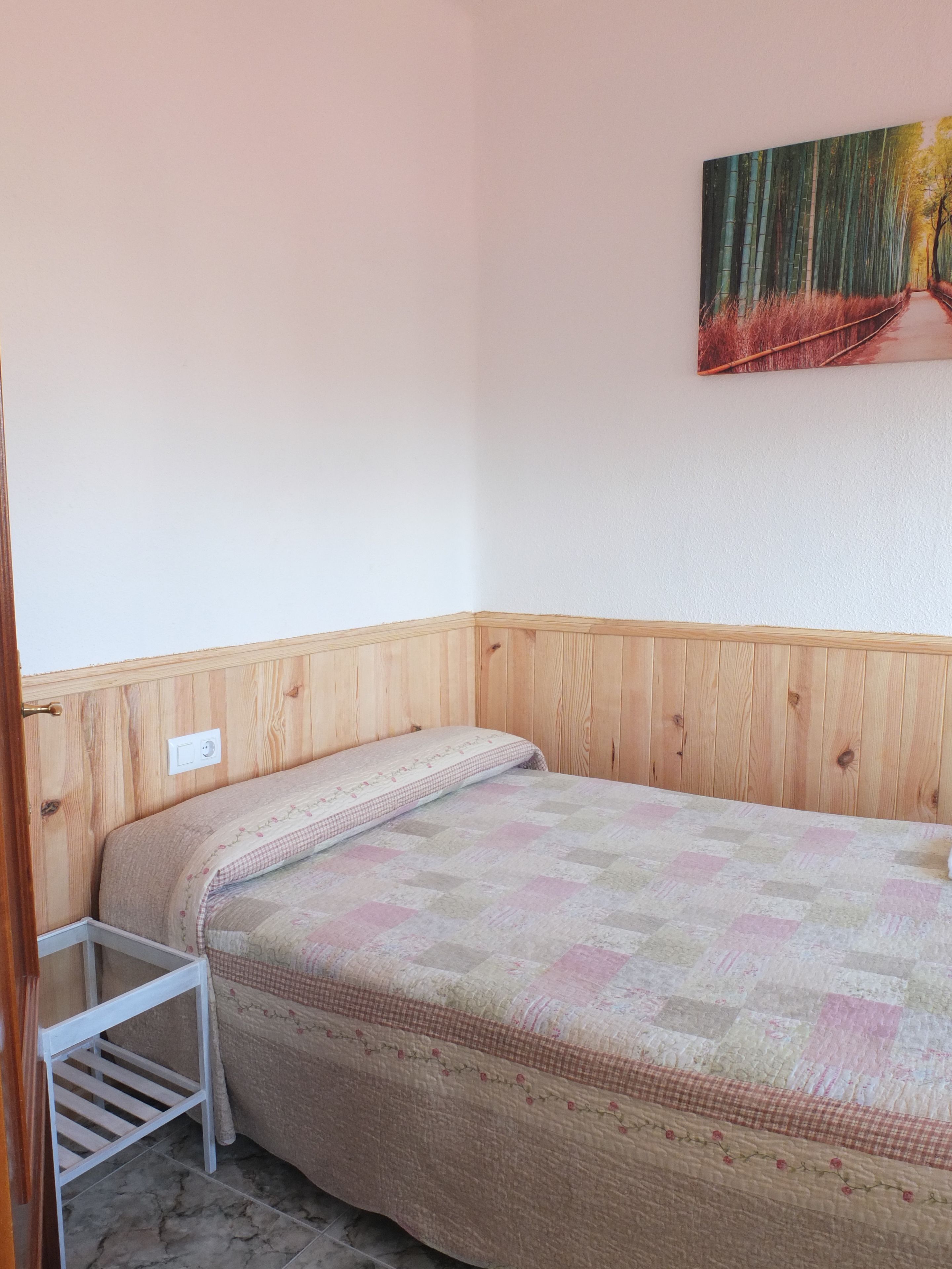 Een kleine, verzorgde kamer met een enkel bed, een houten hoofdbord, een klein plankje en een schilderij aan de muur.