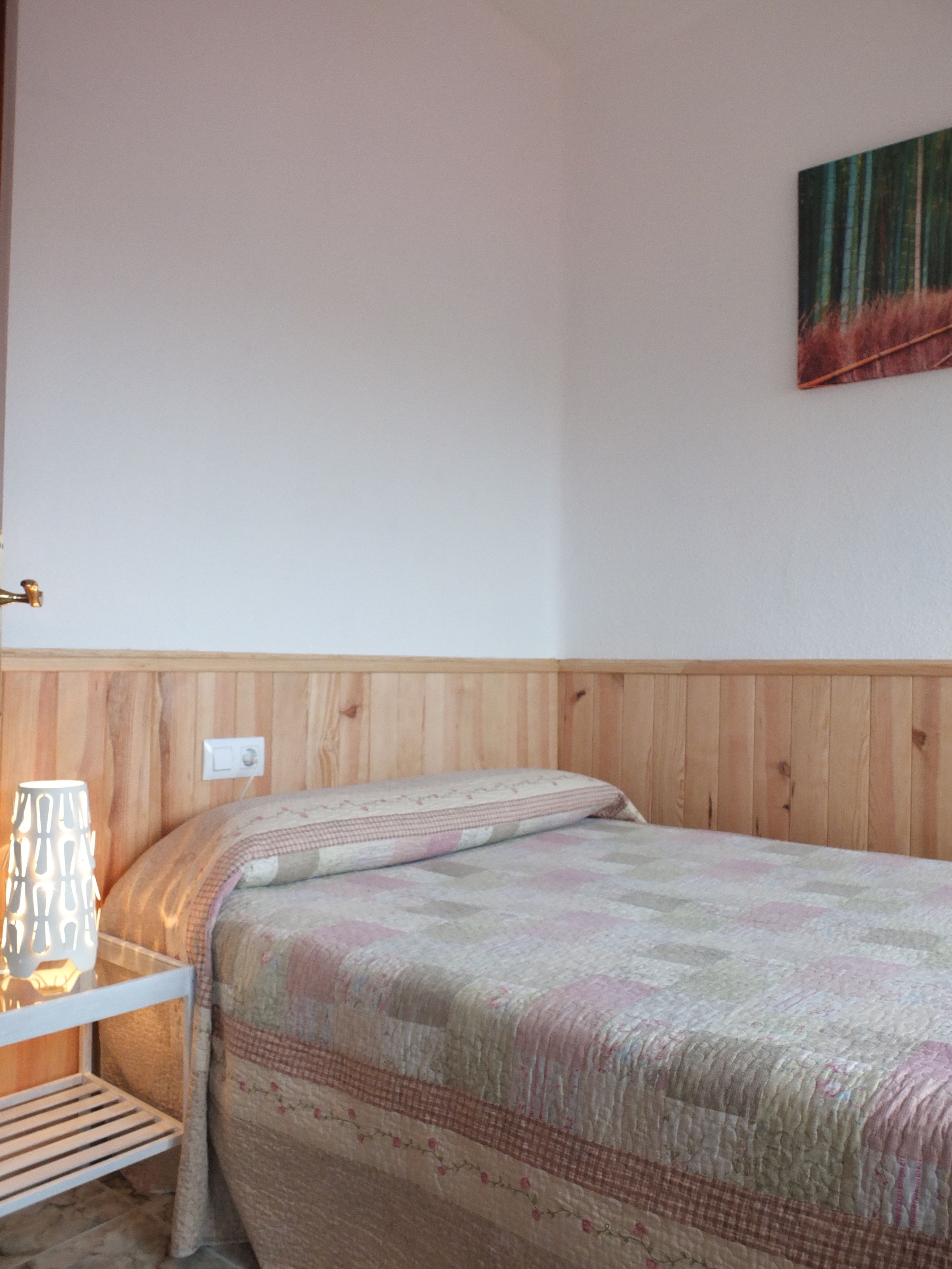 Un dormitorio con cabecera de madera, cama con colcha estampada, mesita de noche con lámpara y un cuadro colgado en la pared.
