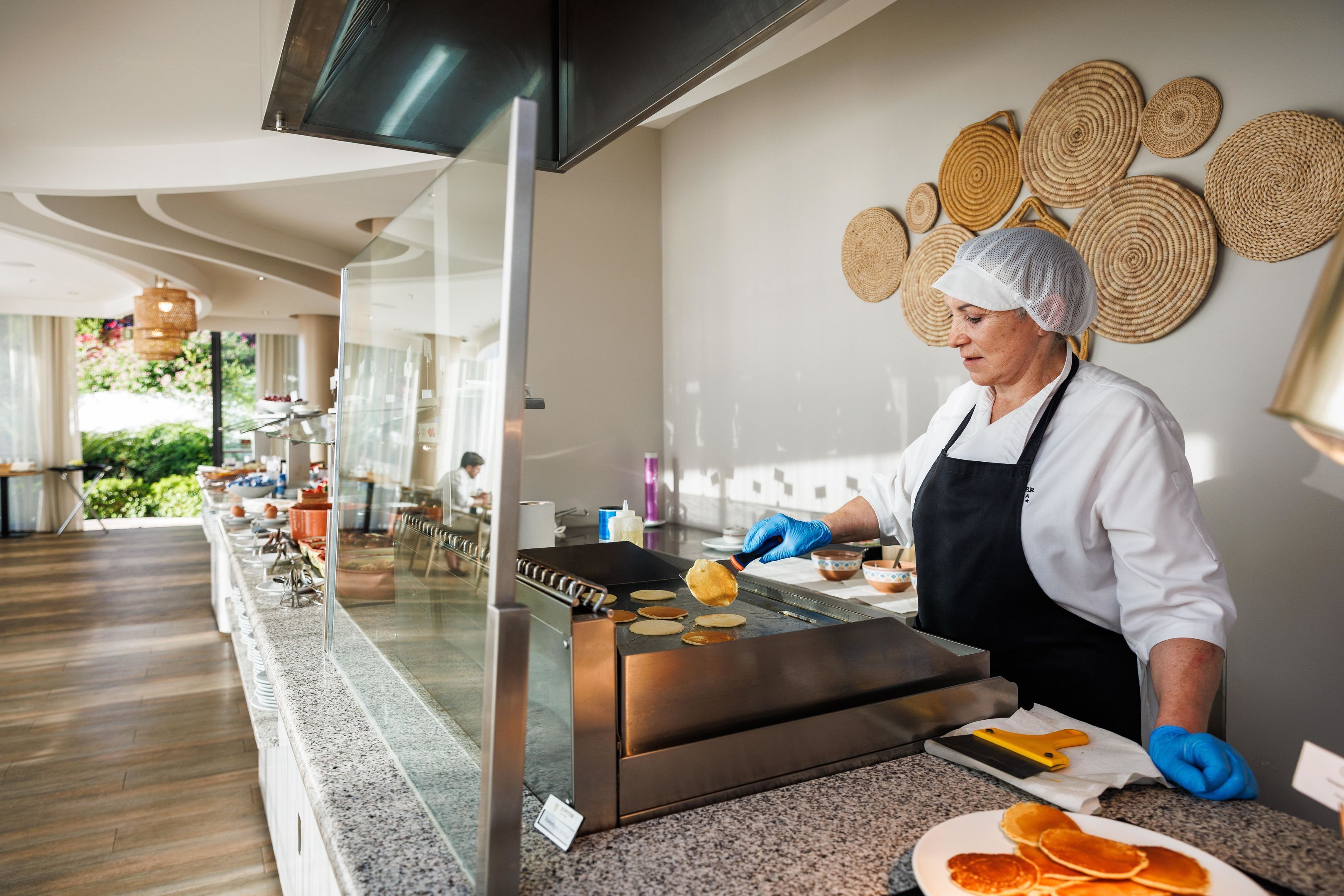 A person cooking pancakes on a griddle in a modern kitchen with a glass partition.