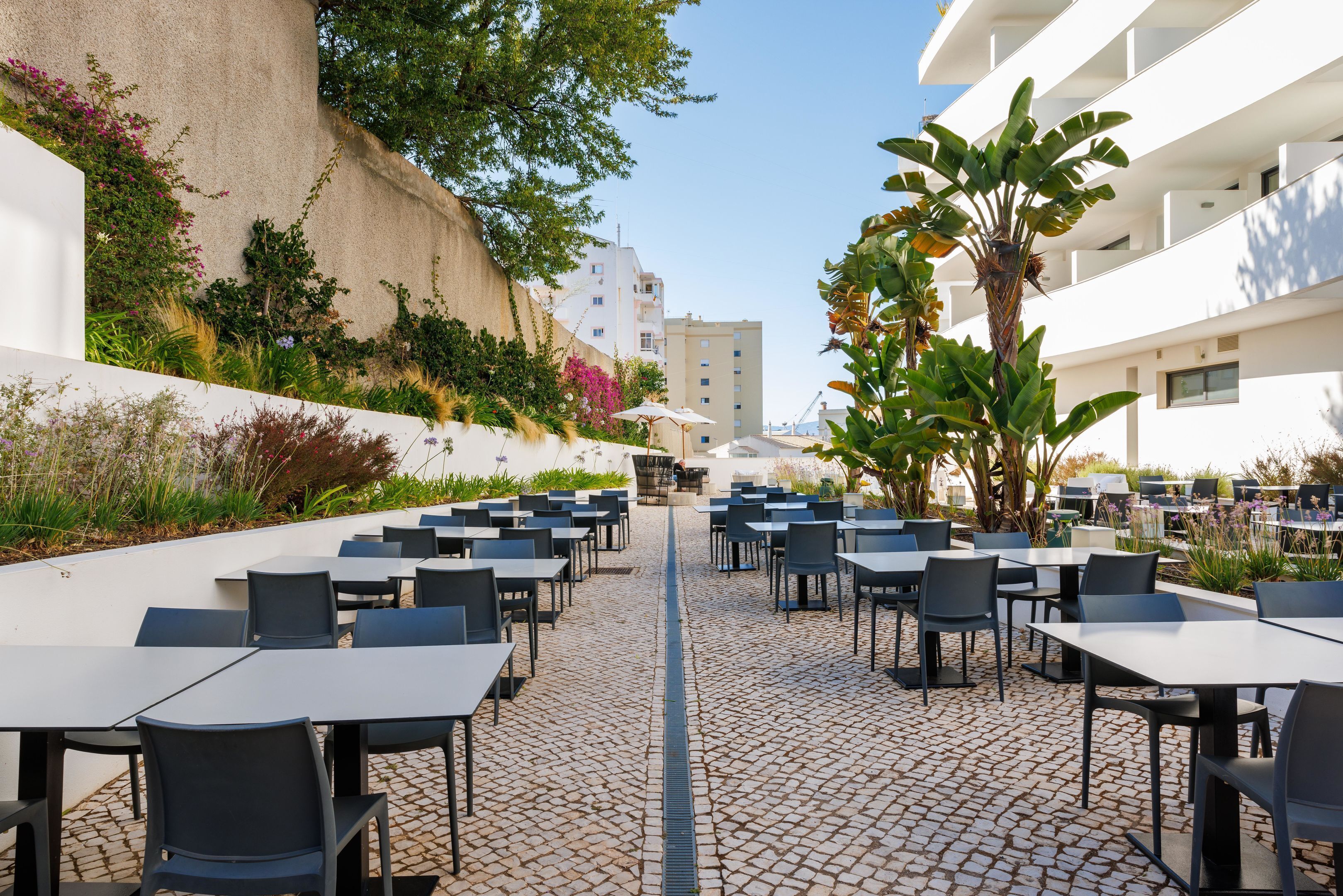 An outdoor dining area with tables and chairs arranged on a cobblestone floor, surrounded by greenery and a building in the background.