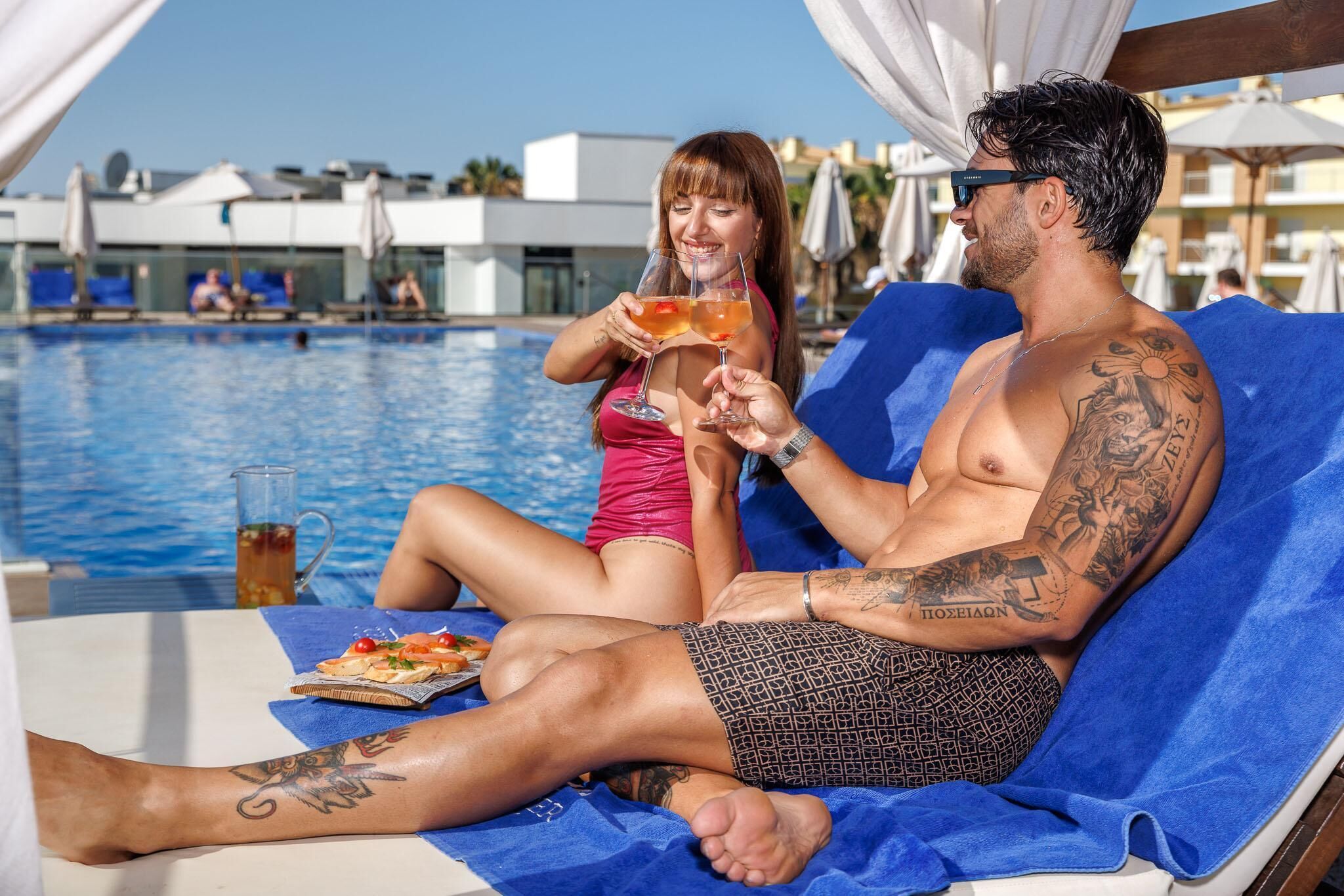A man and a woman relaxing by a poolside lounge chair with a drink and a plate of food.