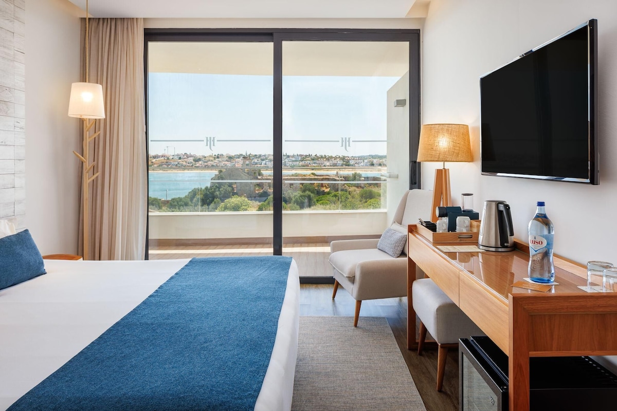 A hotel room with a bed, a desk, a TV, a view of the sea, and a cityscape.