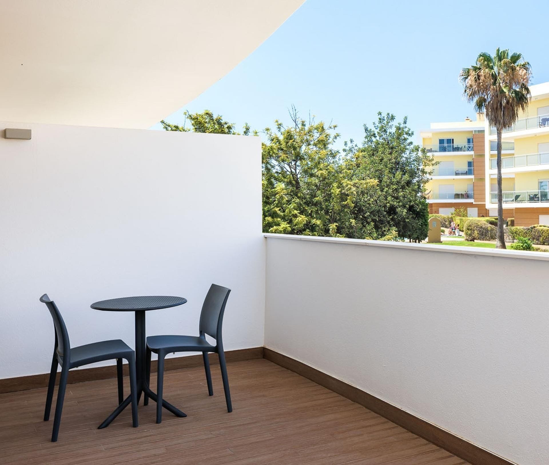Een balkon met een ronde tafel en twee stoelen, een houten vloer en uitzicht op een gebouw en bomen.