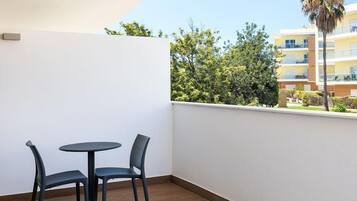 Een balkon met een ronde tafel en twee stoelen, een houten vloer en uitzicht op een gebouw en bomen.
