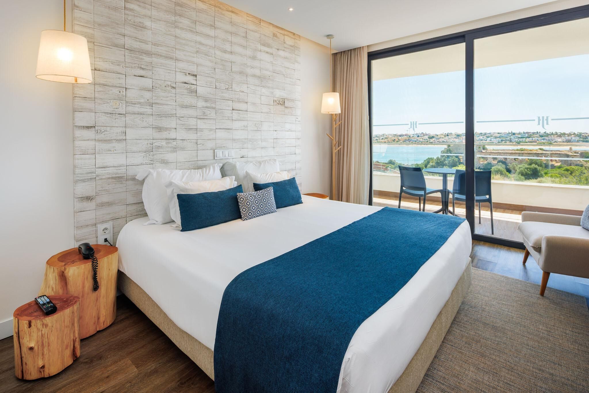 A modern hotel room with a large bed, a wooden headboard, and a balcony with a view of the city and a bridge.
