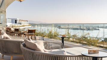 A terrace with wicker furniture, a table with a block number, and a view of a body of water and cityscape.