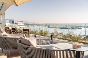 A terrace with wicker furniture, a table with a block number, and a view of a body of water and cityscape. - Jupiter Marina Hotel - Couples & Spa (Portimão)
