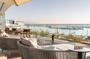 Una terraza con mobiliario de mimbre, una mesa con un número de bloque y vistas a un cuerpo de agua y paisaje urbano.