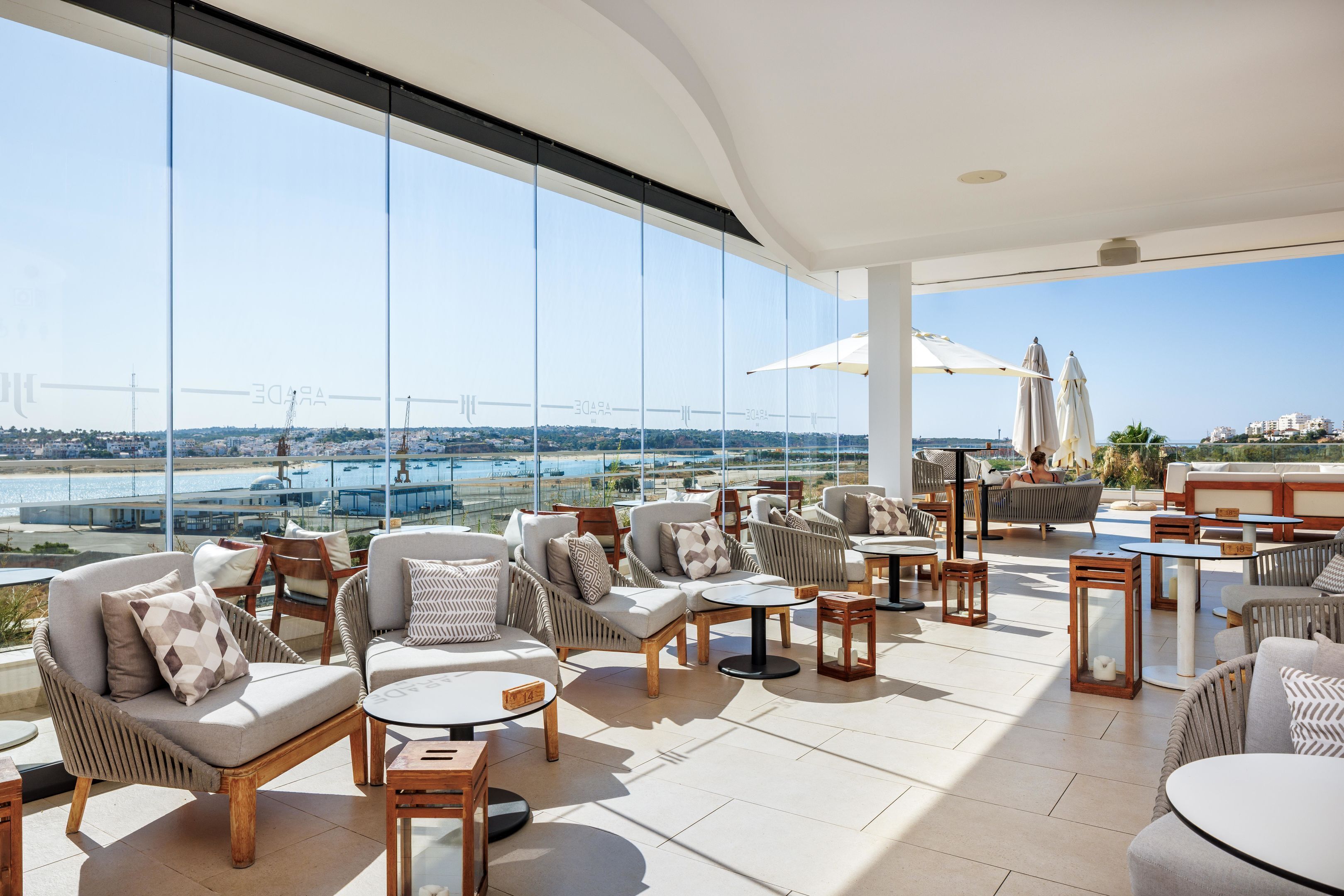 A spacious outdoor lounge area with comfortable seating, large windows offering a view of a bridge and the sea, and a pool in the background.