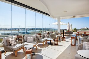 Amplia zona lounge al aire libre con asientos cómodos, ventanas grandes que ofrecen vistas a un puente y al mar, y una piscina al fondo.