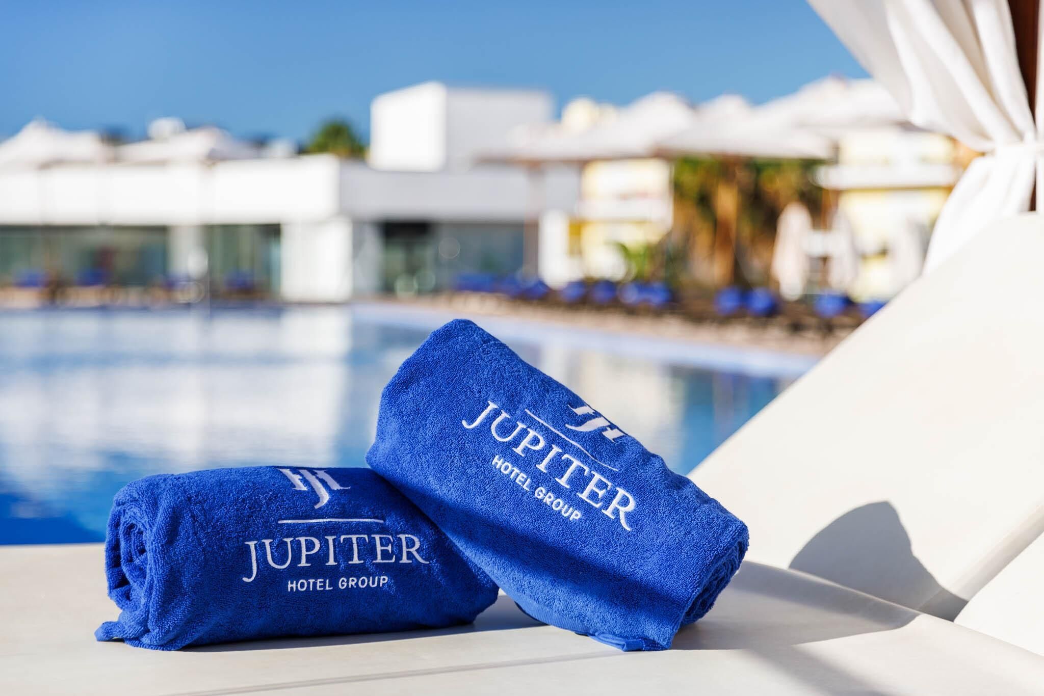 Duas toalhas azuis com a inscrição "JUPITER HOTEL GROUP", colocadas numa cadeira de banho branca junto a uma piscina.