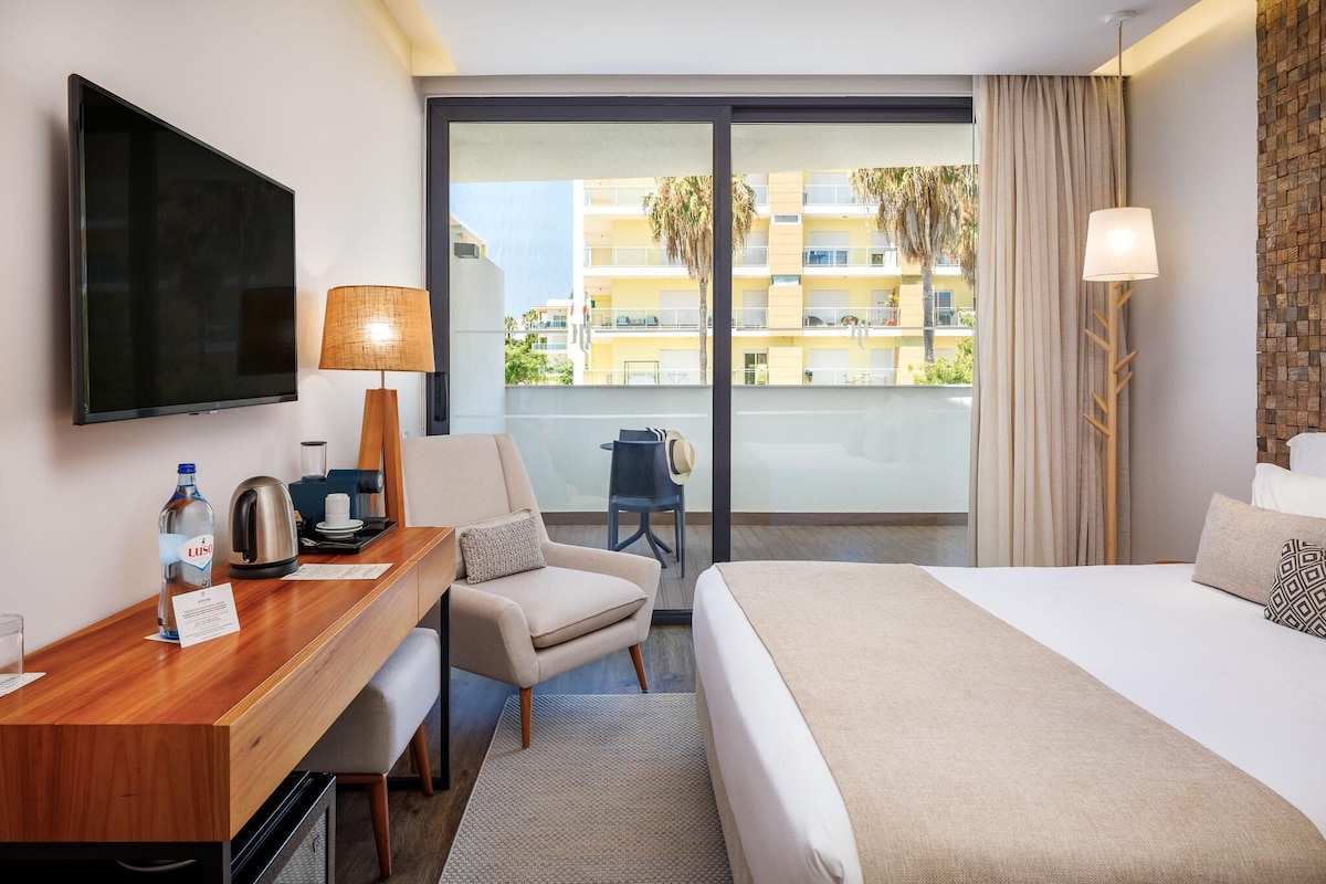 Quarto de hotel com uma cama, uma escrivaninha com luminária, uma televisão e vista para a varanda externa.