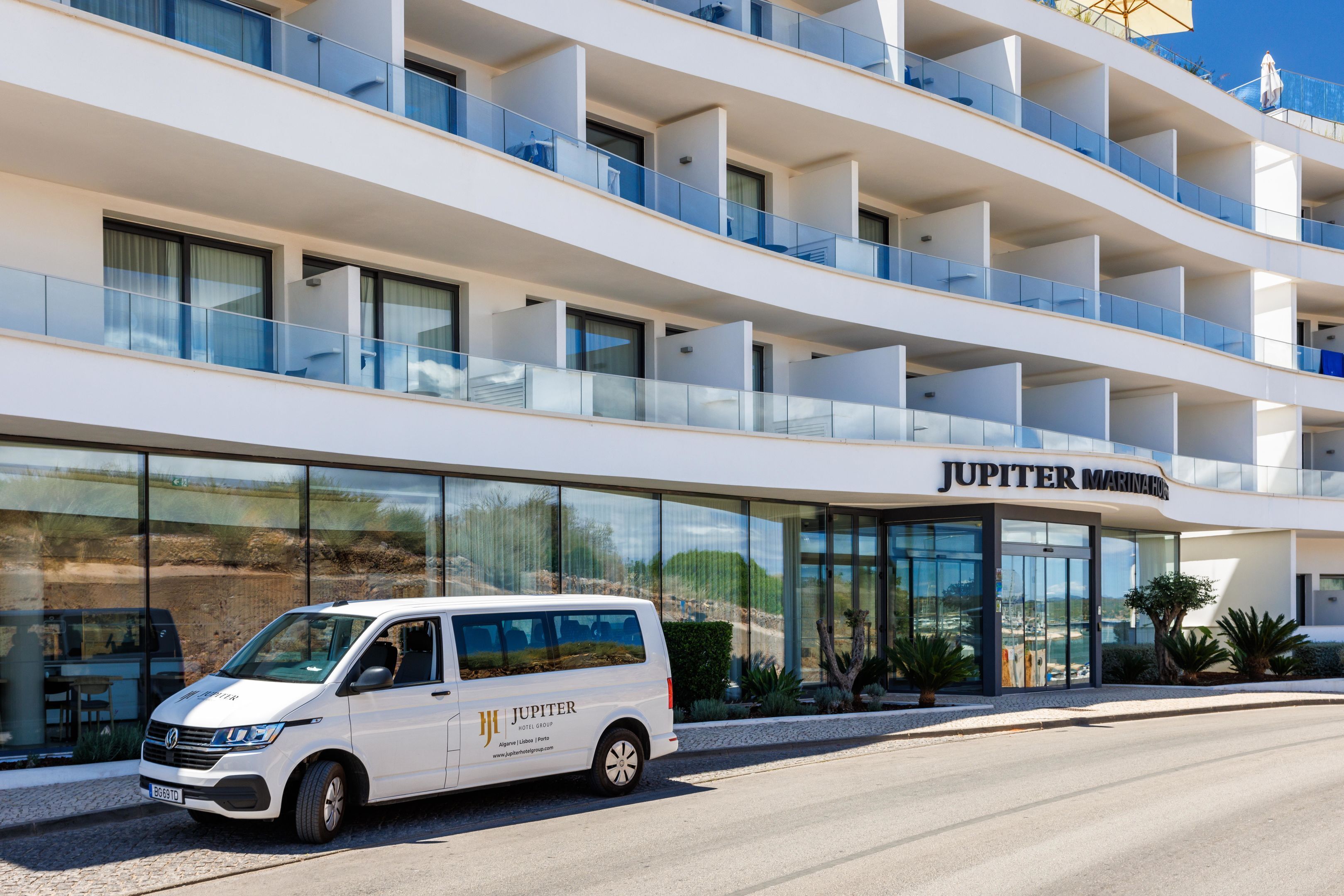 Una camioneta blanca estacionada frente a un hotel moderno con varios balcones y una vista despejada del paisaje circundante.