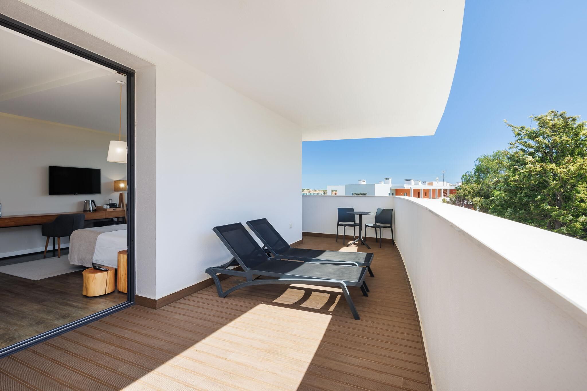Ein Balkon mit zwei Liegestühlen, einem kleinen Tisch und einem Sessel. Im angrenzenden Raum sind ein Fernseher und ein Schreibtisch mit Stuhl zu sehen.