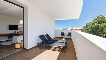 A balcony with two lounge chairs, a small table, and a chair. A television and a desk with a chair are visible in the adjacent room.