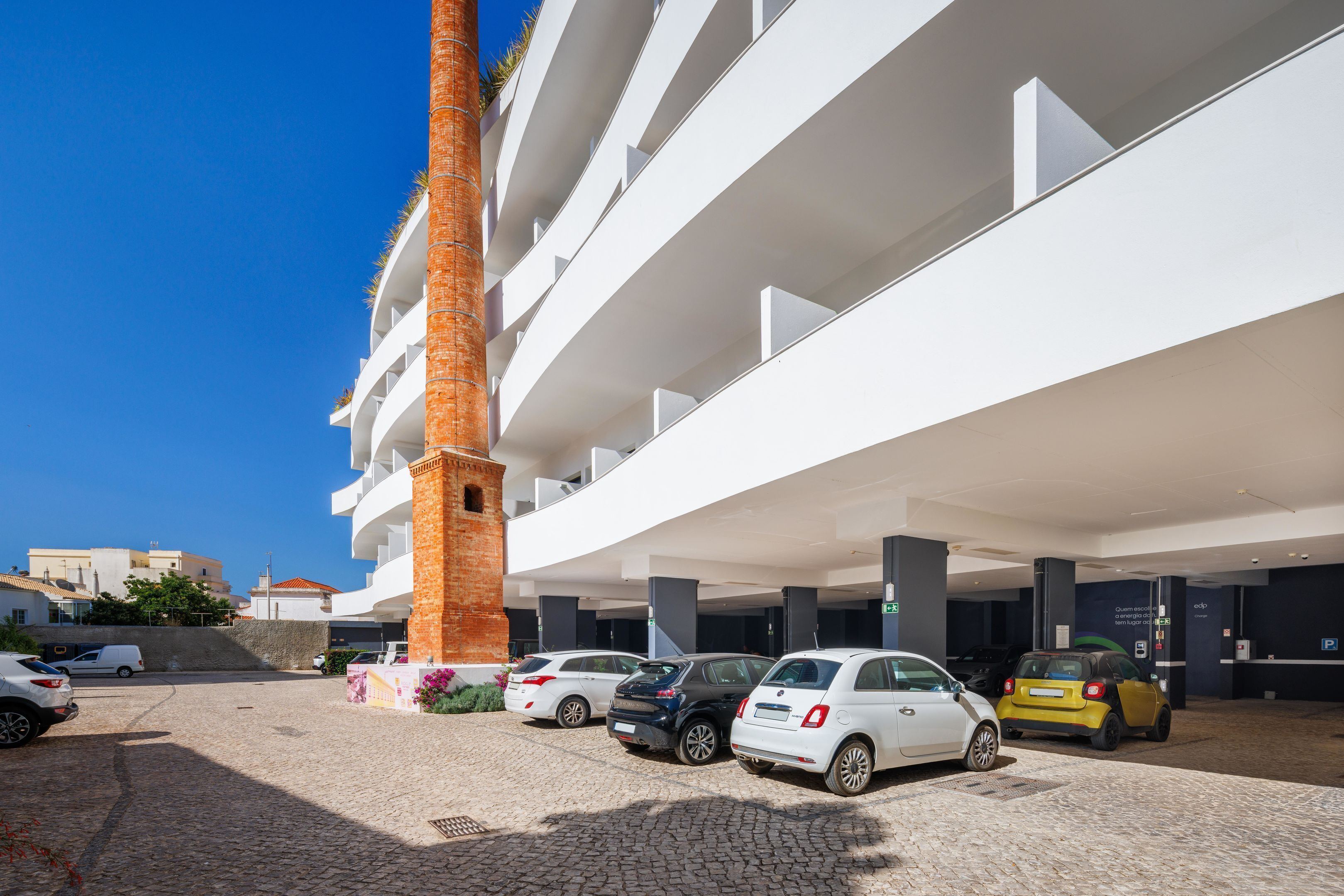 Um edifício moderno com vários andares, uma área de estacionamento com diversos carros e um colunar de tijolos imponente.