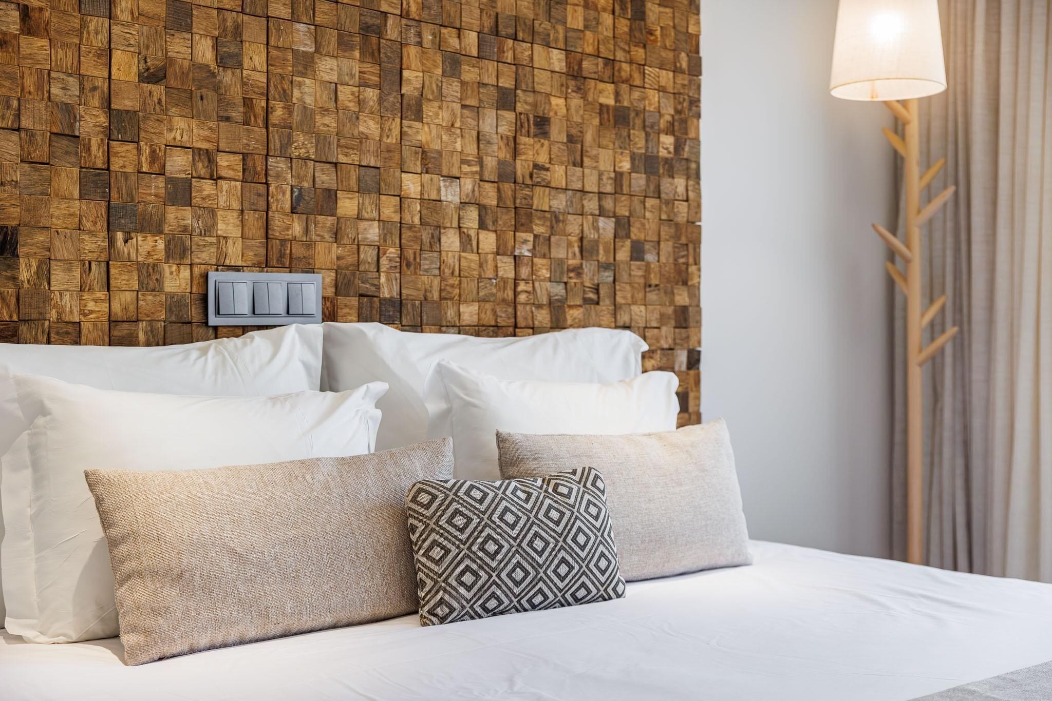 A neatly made bed with white linens and several pillows, a wooden textured wall behind, and a lamp on the right.