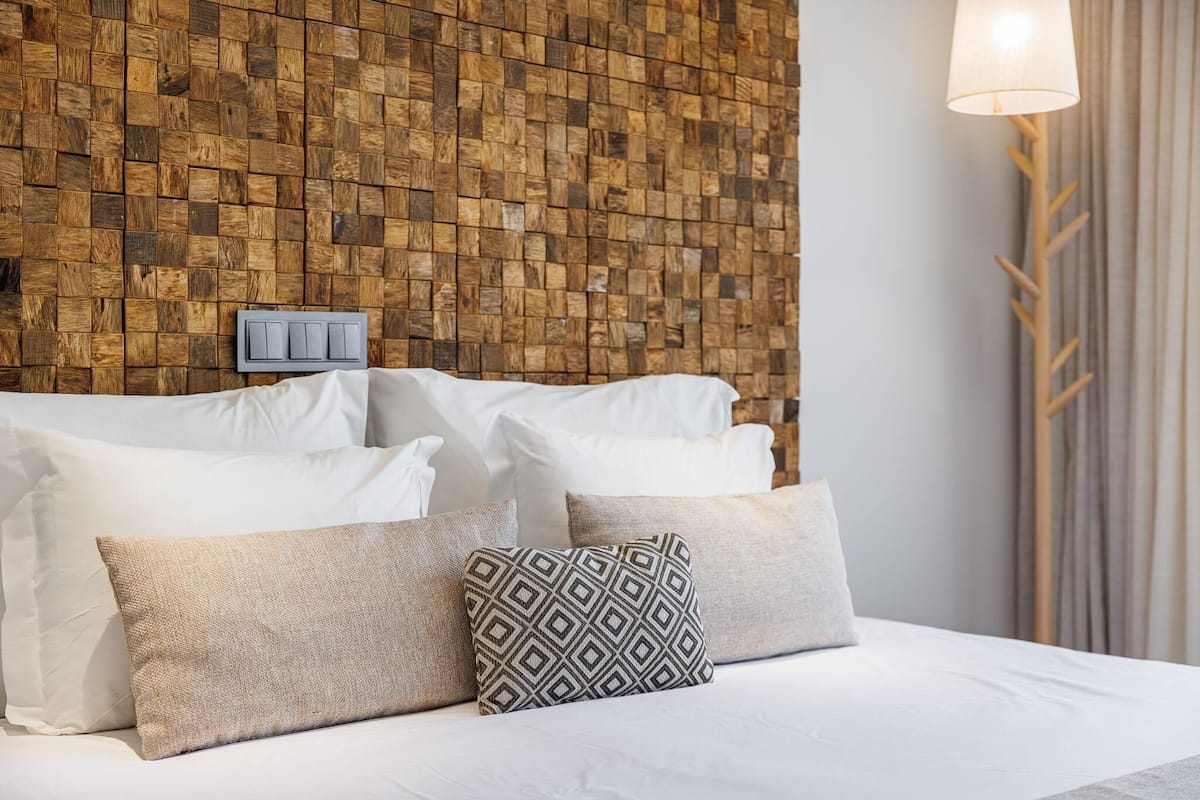 A neatly made bed with white linens and several pillows, a wooden textured wall behind, and a lamp on the right.