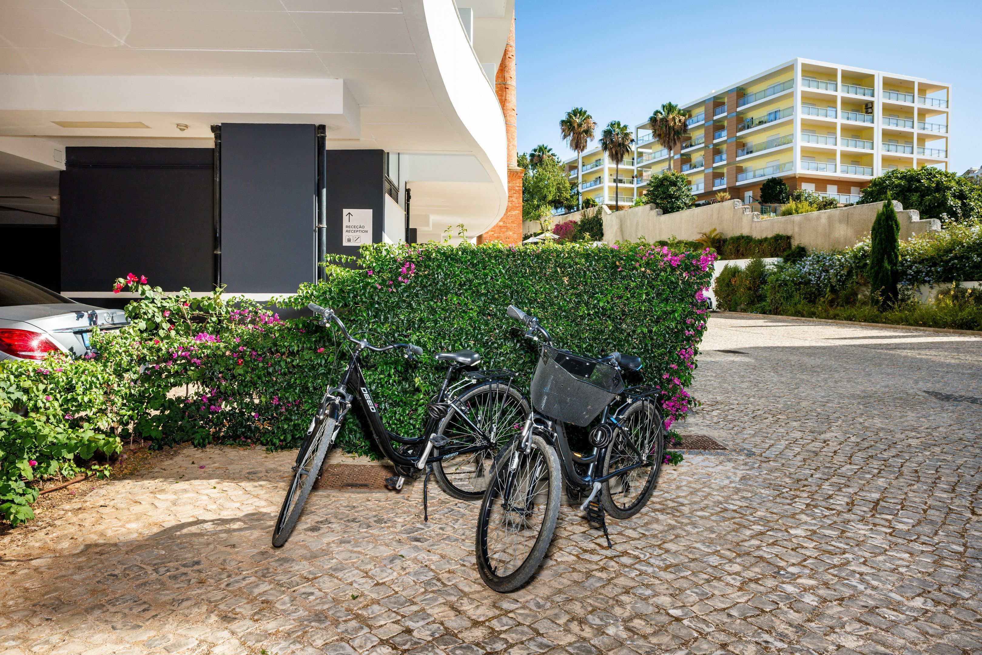 A bicycle parked in front of a building with a modern design, a paved area, and a multi-story structure in the background.