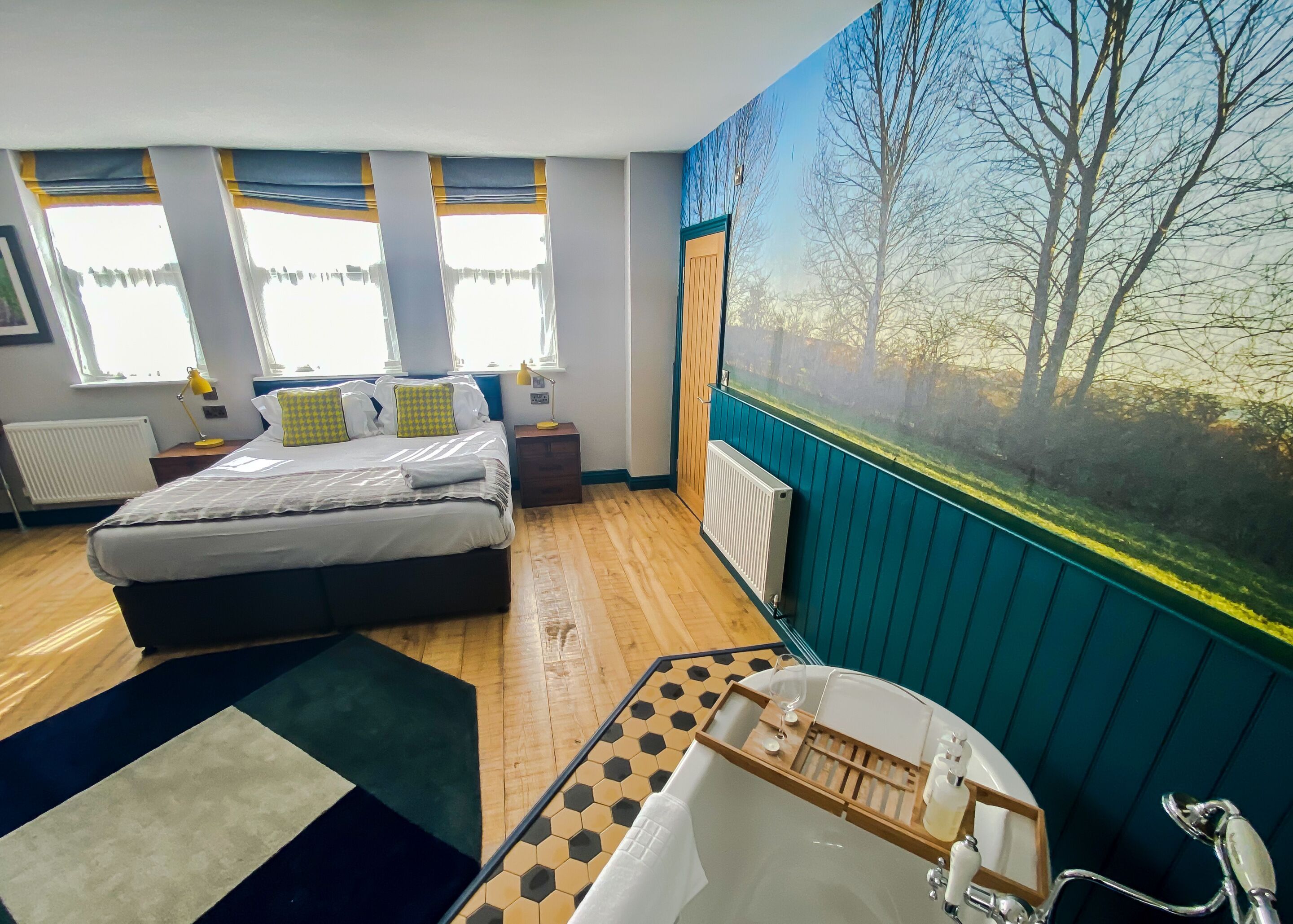 A bedroom with a bed, bedside tables, a window with curtains, a radiator, a large window with a view of trees, and a bathroom with a bathtub and a mirror.