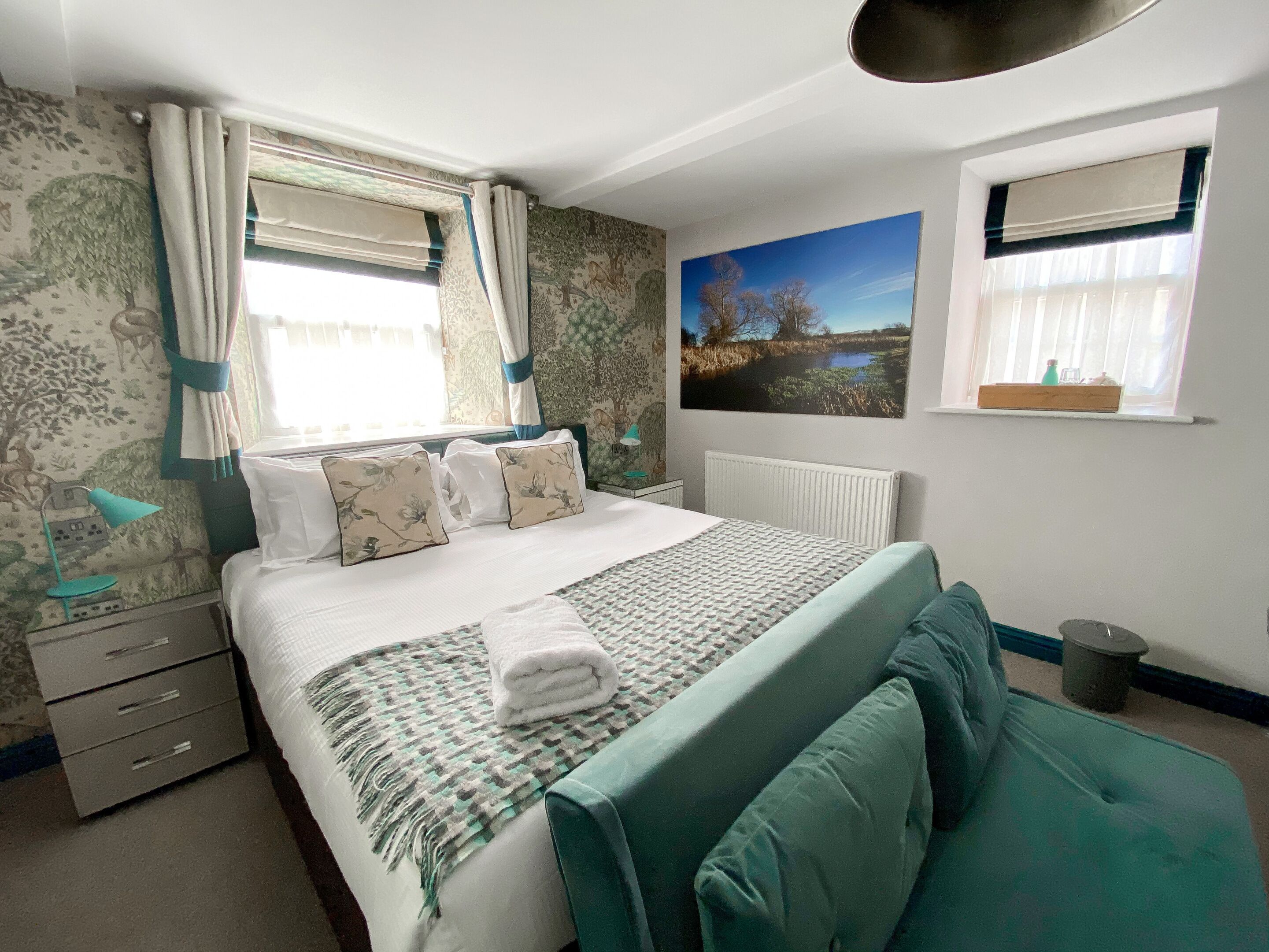 A bedroom with a bed, a bedside table, a lamp, a radiator, a window with curtains, and a wall-mounted picture.