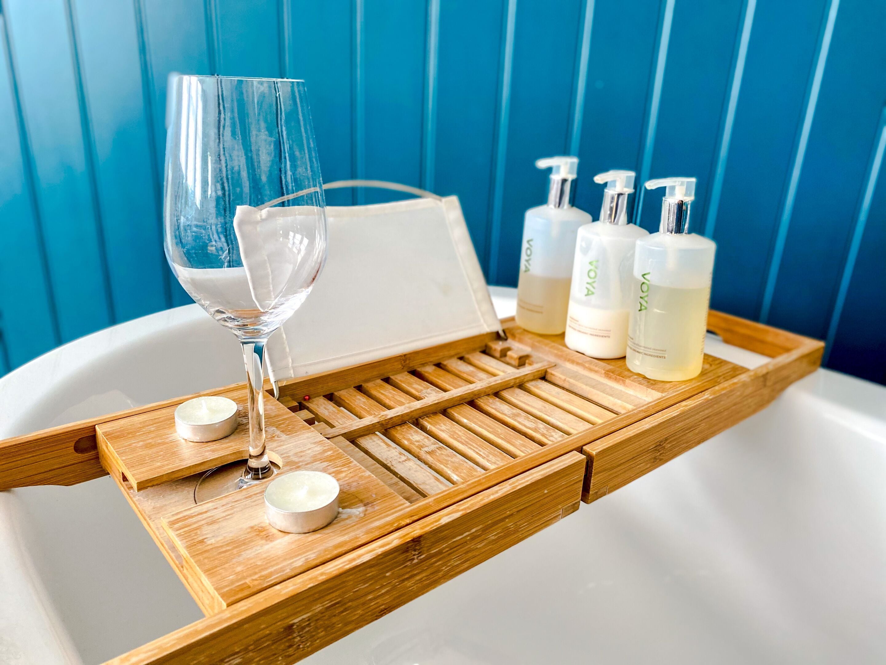 Un vassoio di legno con un bicchiere d'acqua, una candela e tre prodotti Voya su una vasca da bagno.