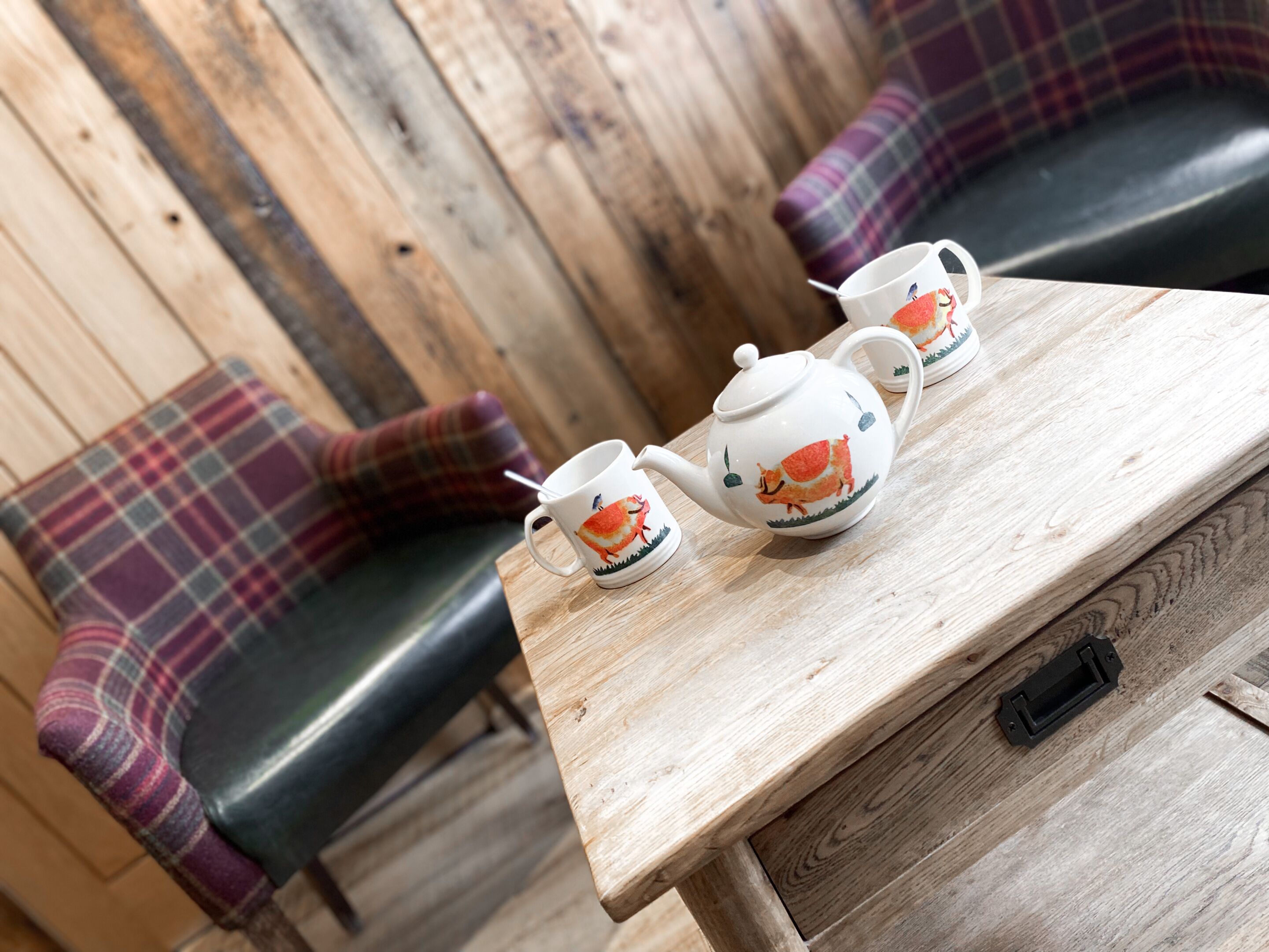 A wooden table with a teapot and two cups, set against a backdrop of plaid-patterned chairs and a wooden wall.