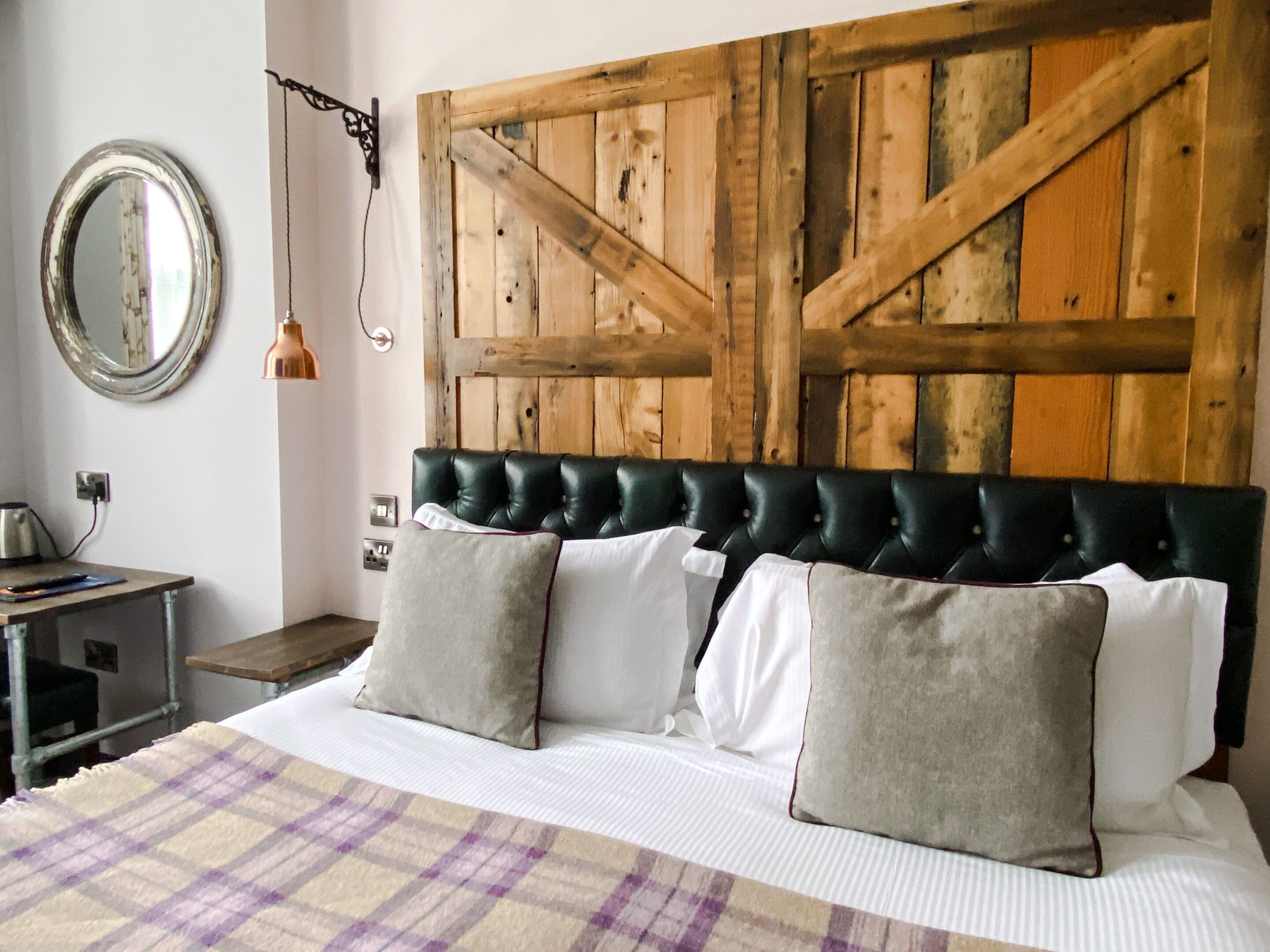 A bedroom with a wooden headboard, a bed with white linens and two pillows, a round mirror, and a small table with a lamp.