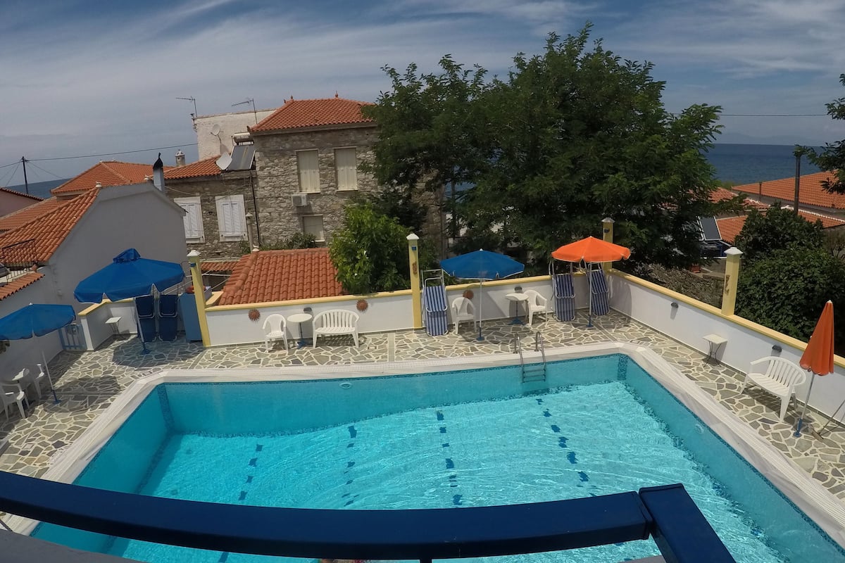 A swimming pool with a stone-tiled deck, surrounded by white chairs and umbrellas, with a view of a building with a red-tiled roof and a tree in the background.