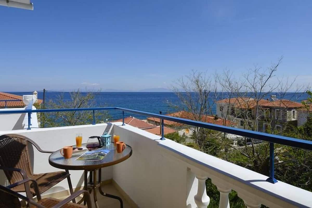 A balcony with a table and chairs, overlooking a coastal view with buildings and the sea.