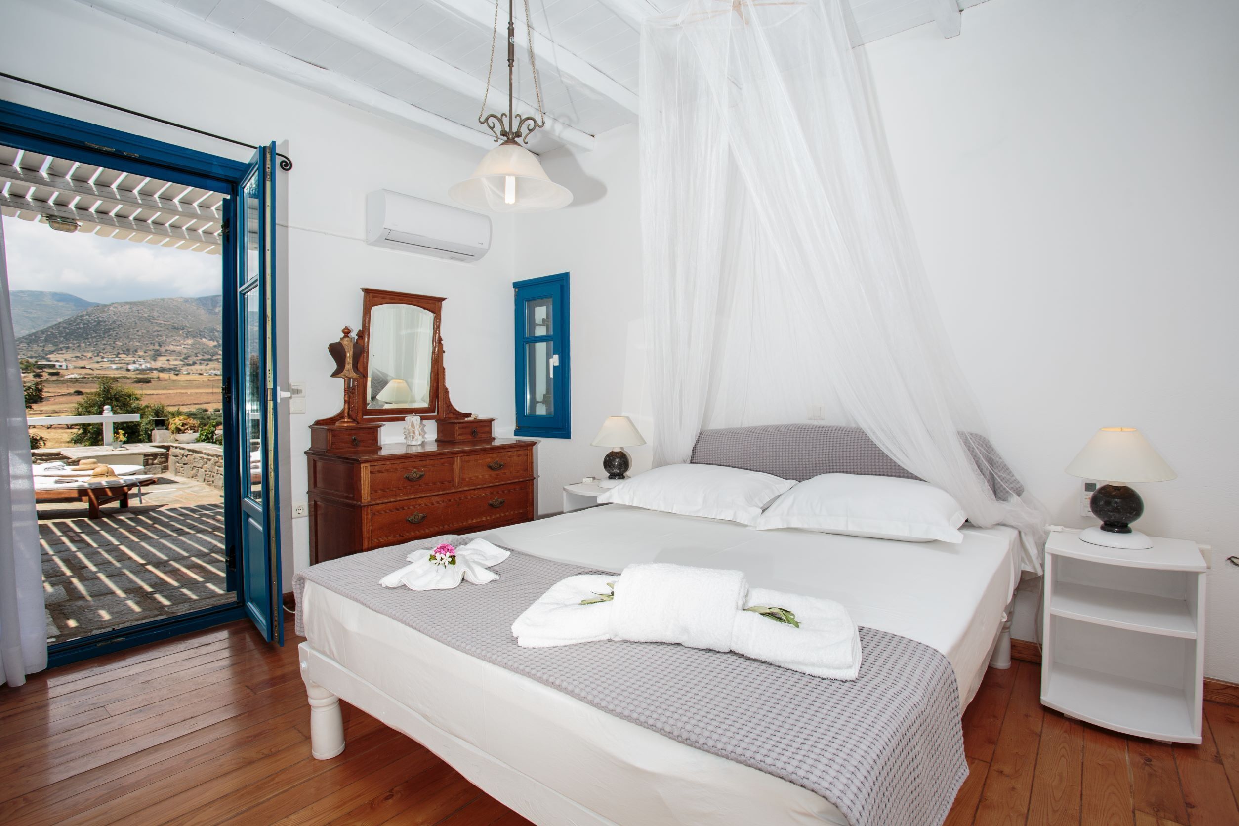 A bedroom with a large bed, a wooden dresser, a nightstand, and a view of the outdoors through an open door.