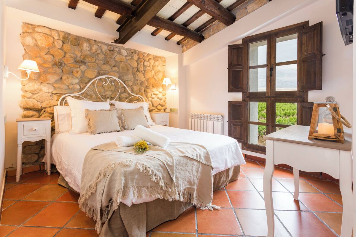 A bedroom with a stone wall, a bed with white bedding, a nightstand, a window with wooden shutters, and a small table with a candle.