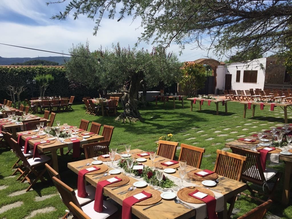 An outdoor dining area with multiple tables set for a meal, surrounded by greenery and a white building in the background.