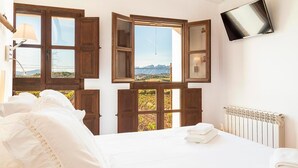 A bedroom with a bed, a TV mounted on the wall, and multiple windows with wooden frames. - Masia Can Canyes & Spa (Sant Llorenc d'Hortons)