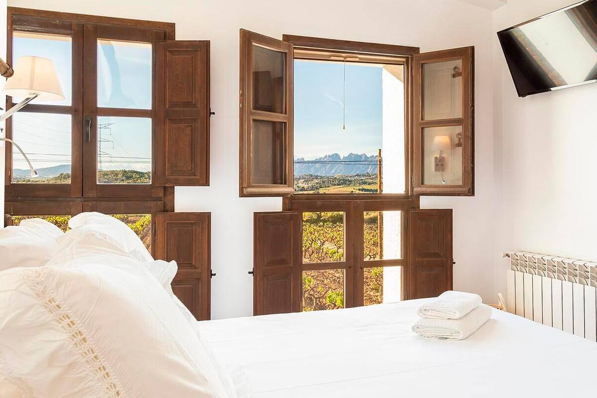 A bedroom with a bed, a TV mounted on the wall, and multiple windows with wooden frames.
