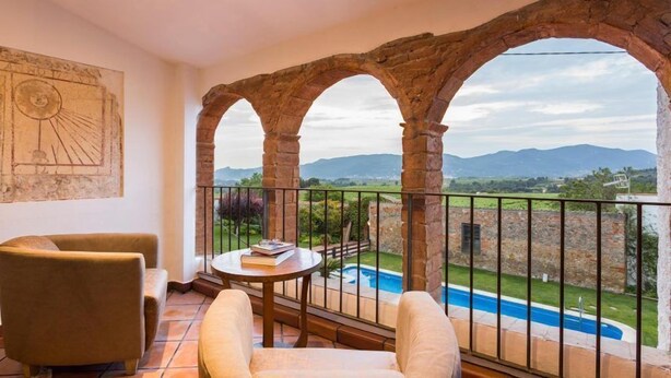 A balcony with a view of mountains, a pool, and a table with books. - Masia Can Canyes & Spa (Sant Llorenc d'Hortons)