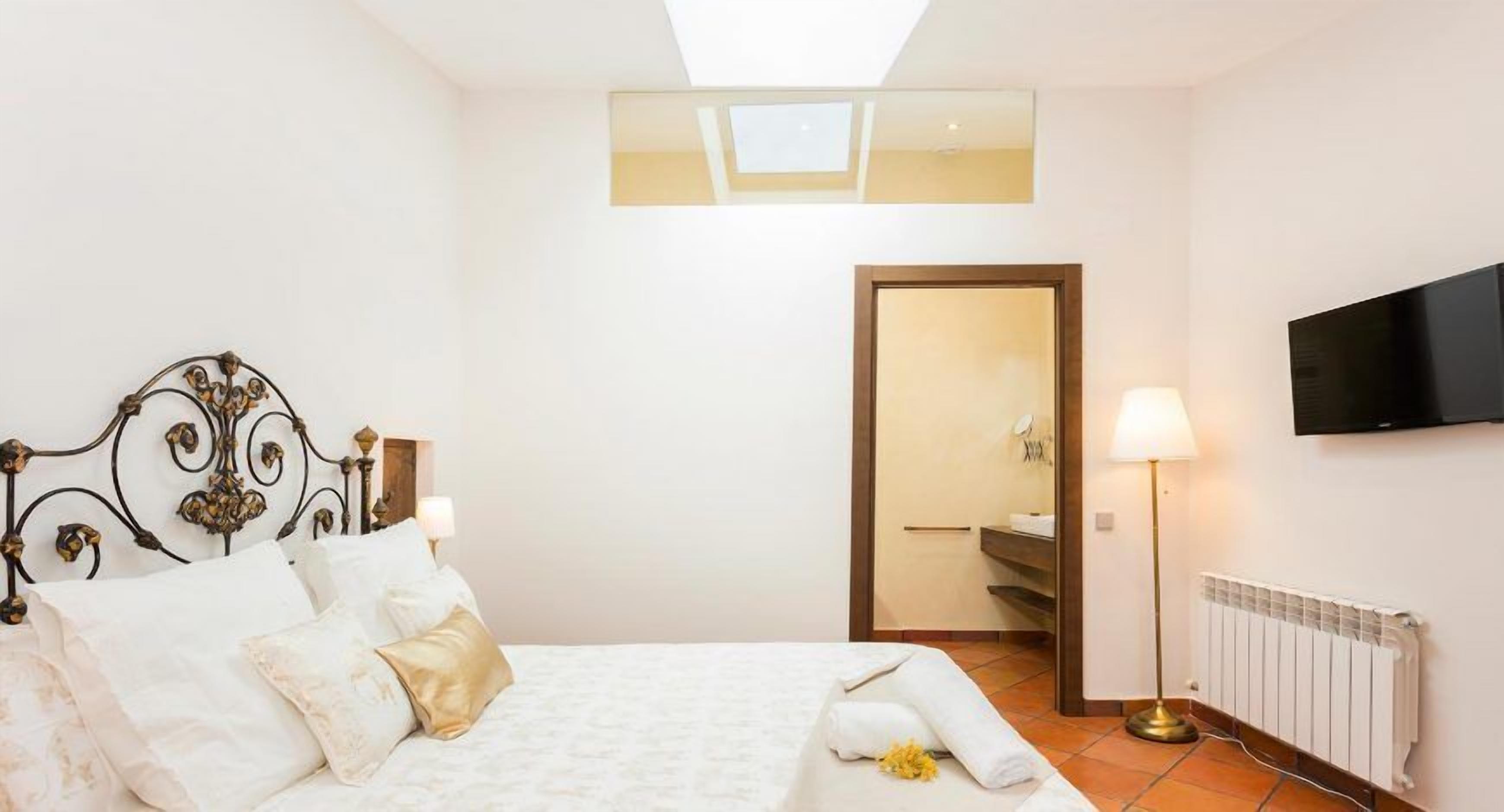Habitación de hotel con cama, lámpara de noche, televisor y vista al baño por una puerta abierta.