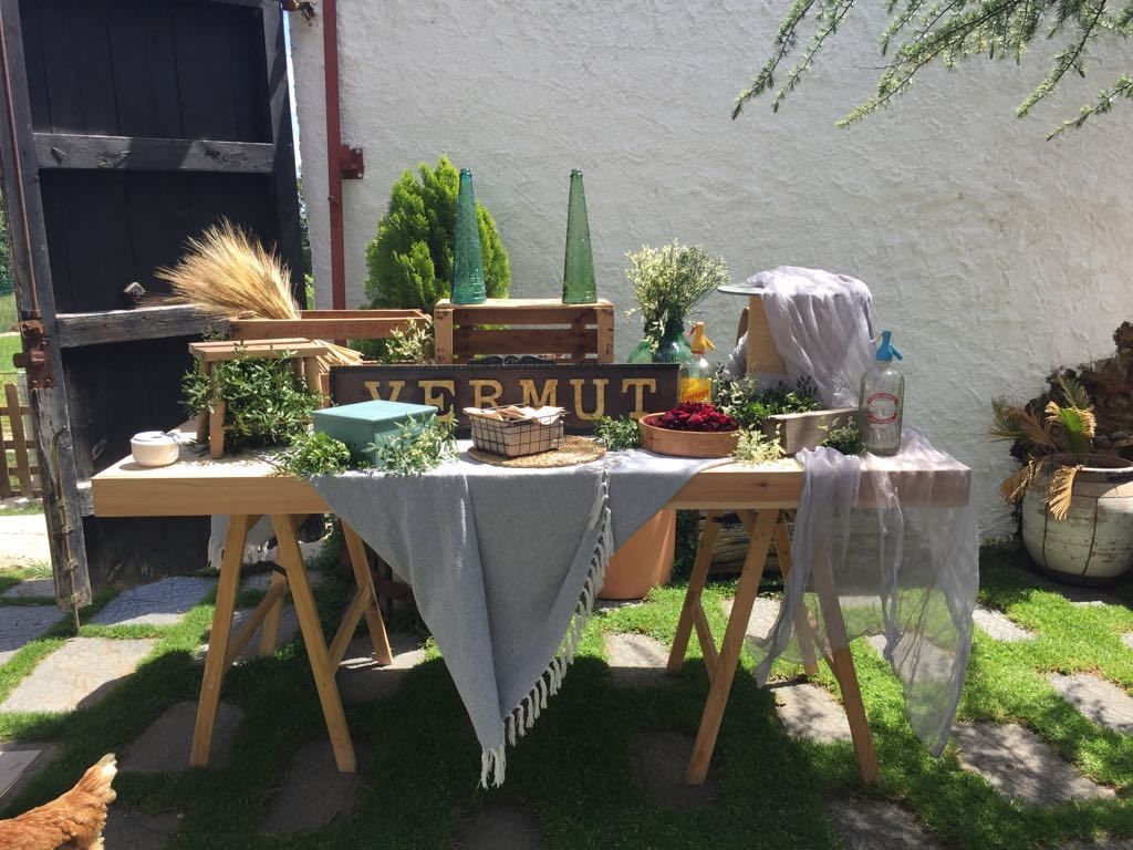 Un área de comedor al aire libre con una mesa de madera dispuesta para dos personas, rodeada de plantas en macetas y un letrero que dice "VERMUT".