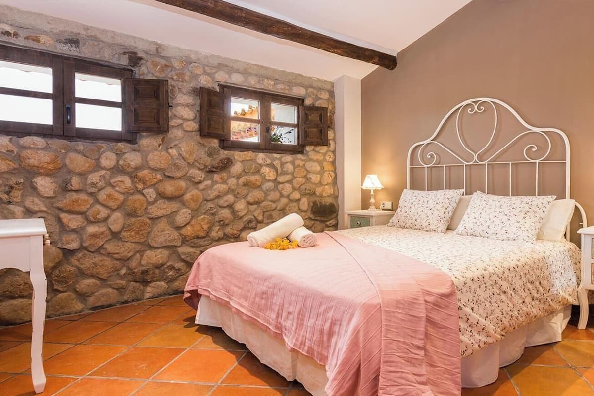 A bedroom with a stone wall, a bed with a white metal headboard, two bedside tables with lamps, and a small white nightstand.