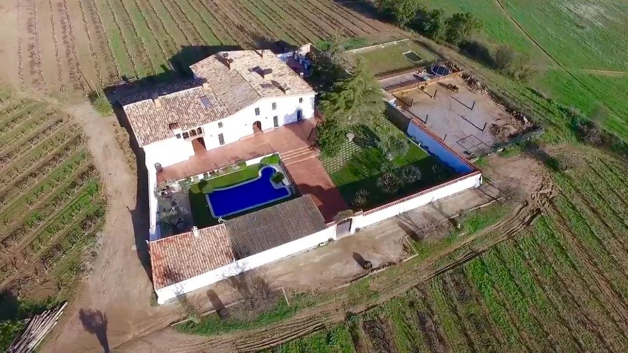 Vista aérea de una extensa propiedad con piscina, cancha de tenis y un edificio blanco de techo con tejas.