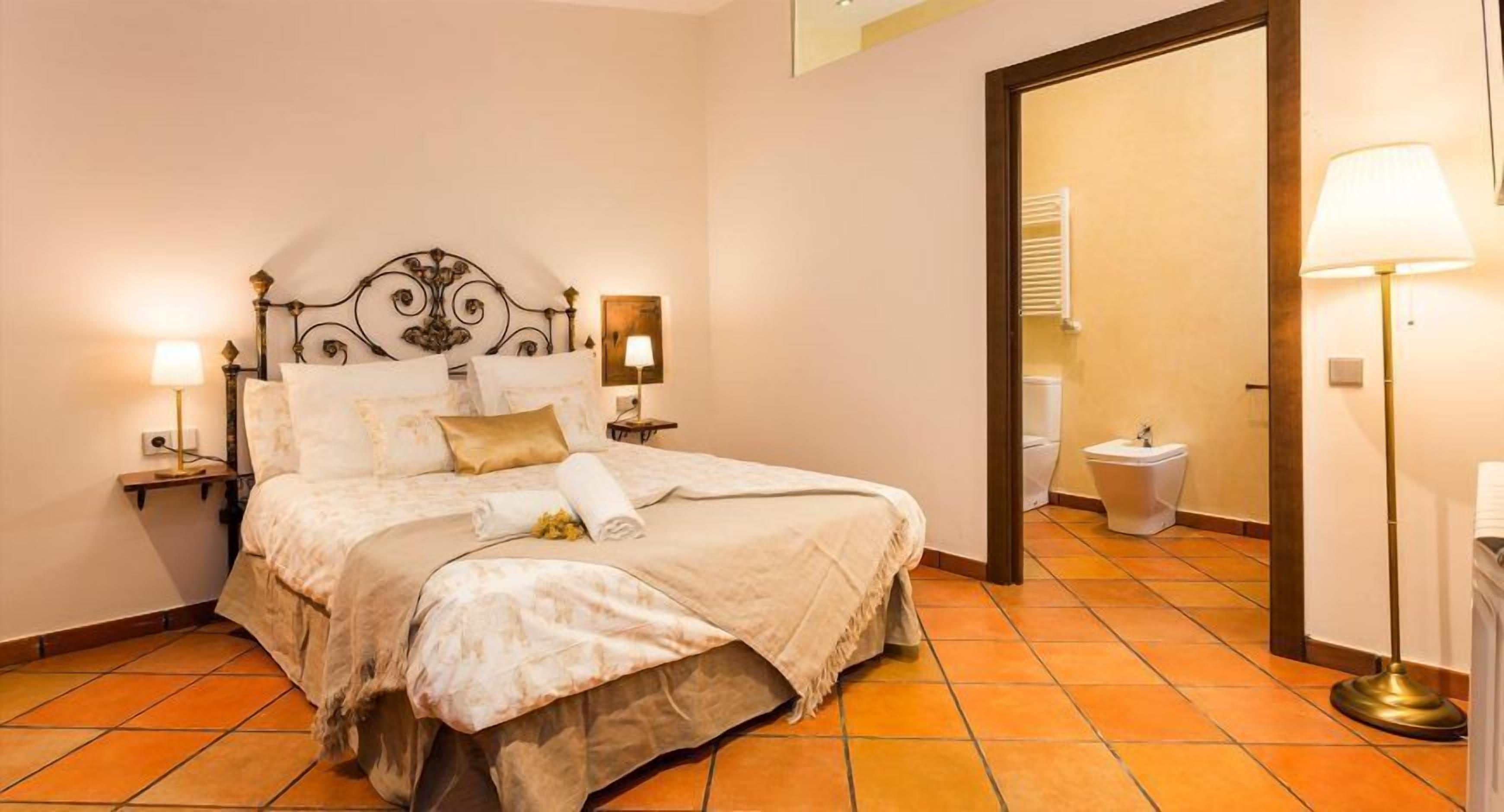 Un llit ben fet amb un capçal, dues làmpades de nit i un mirall que reflecteix un bany amb un vàter i un bidet.