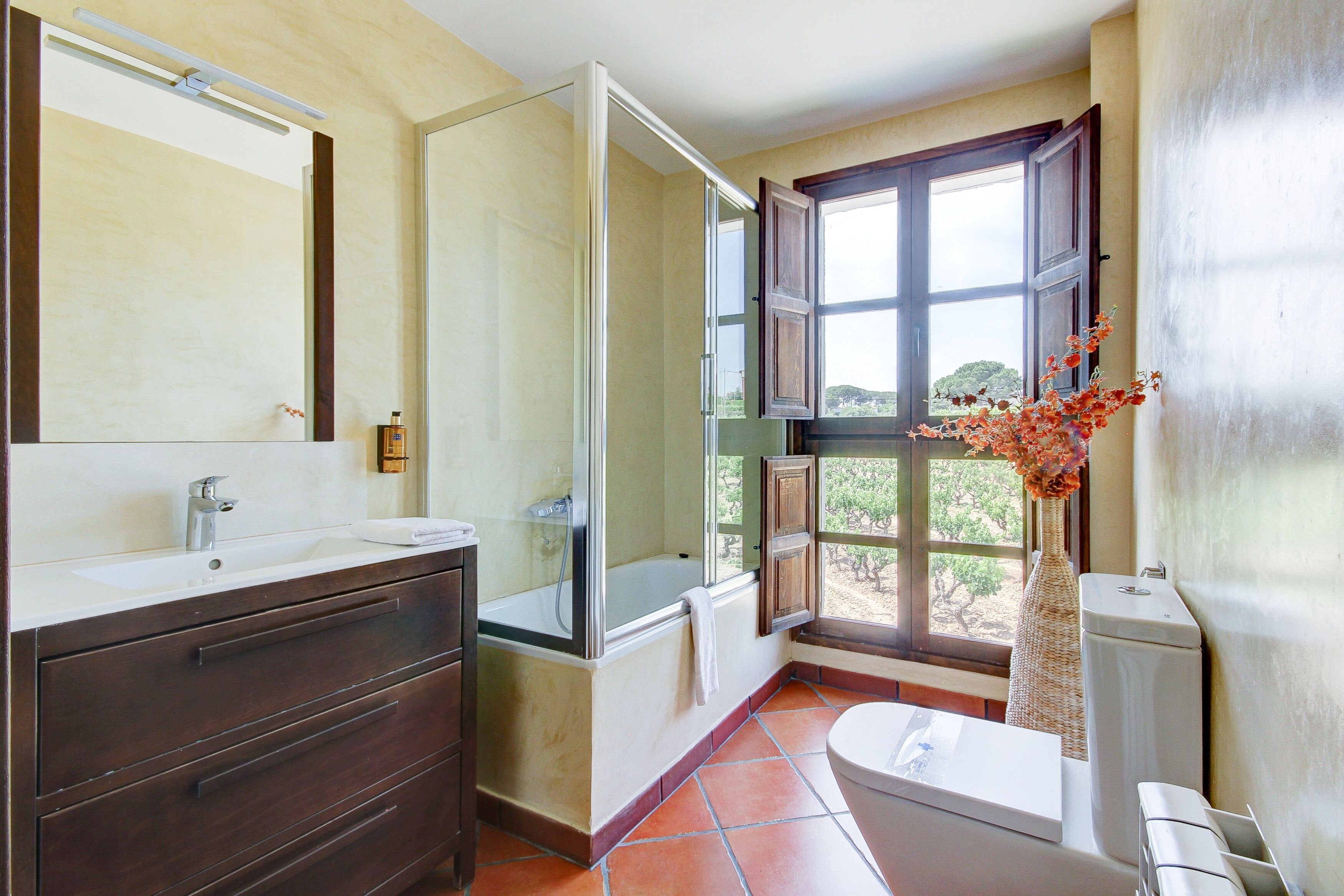 A bathroom with a large mirror, a bathtub, a toilet, and a window with a view of trees.