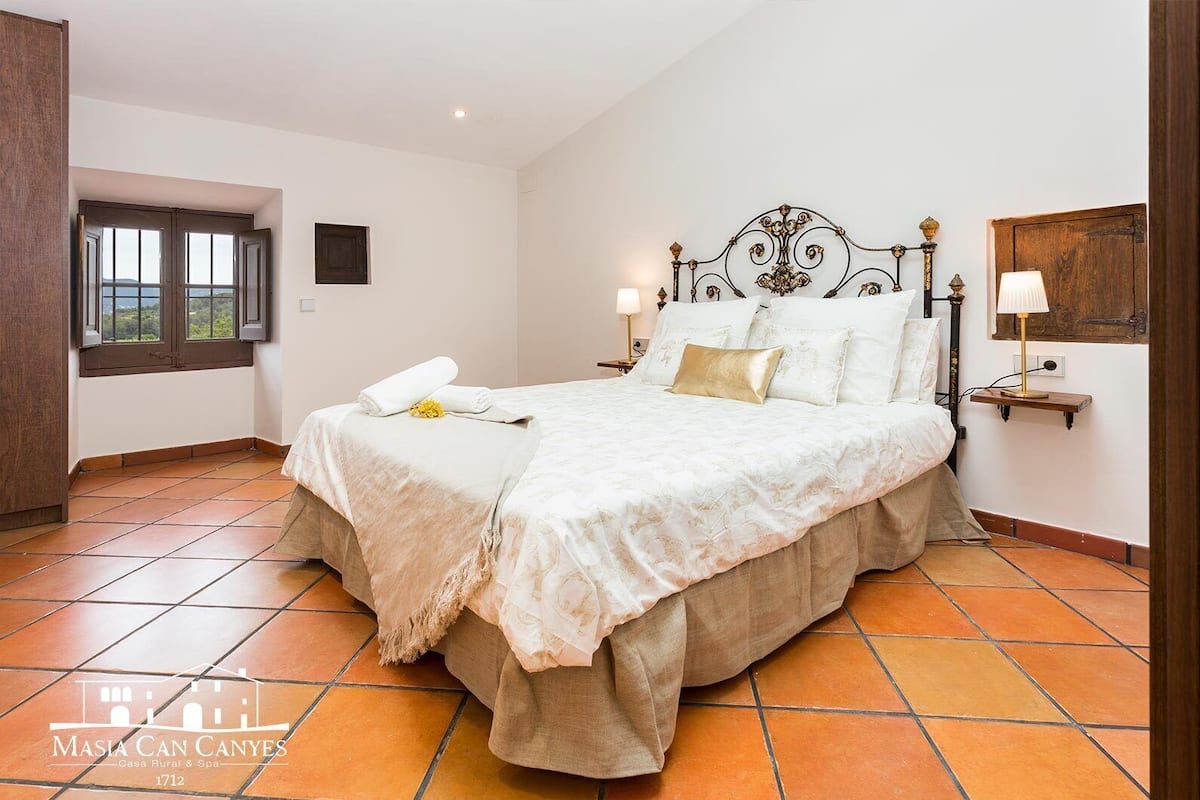A bedroom with a bed, a nightstand, a wall-mounted lamp, a window, and a door.
