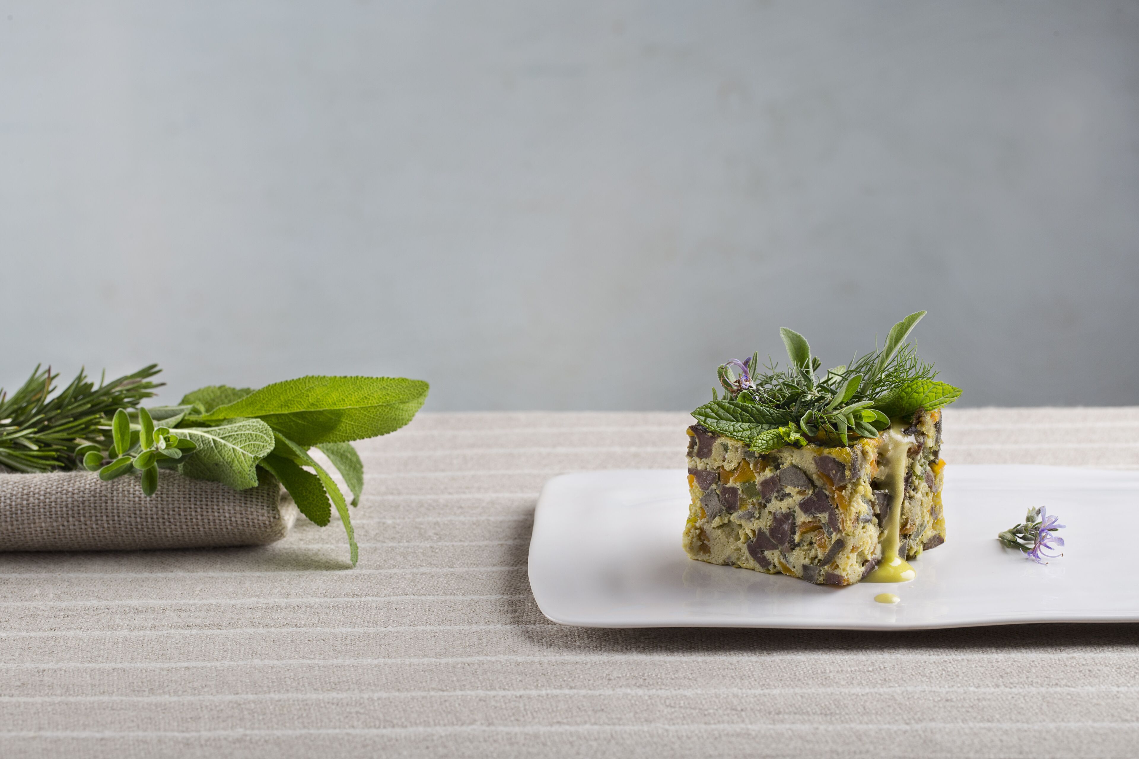 A square white plate with a square food item garnished with herbs, placed on a textured surface.