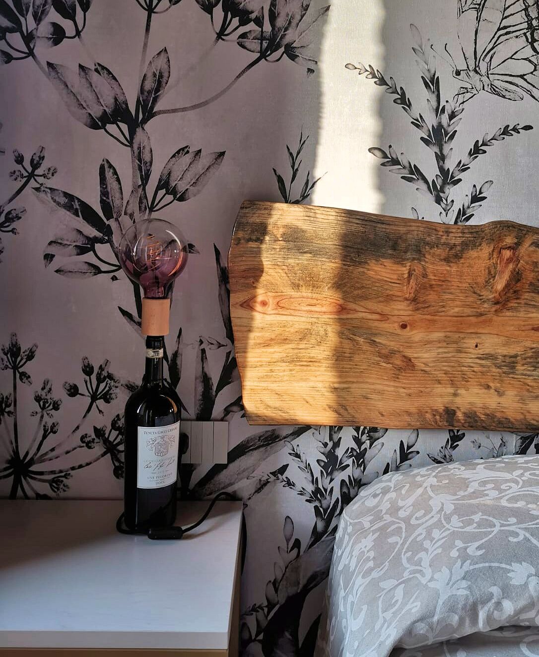 Une tête de lit en bois se détache sur un fond de papier peint à motifs floraux, et une bouteille de vin est posée sur une table de chevet.