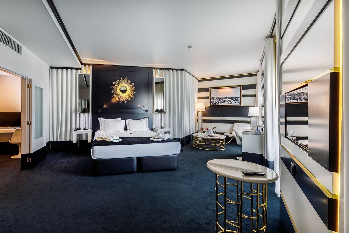 A modern hotel room with a large bed, a round coffee table, and a small bar area.