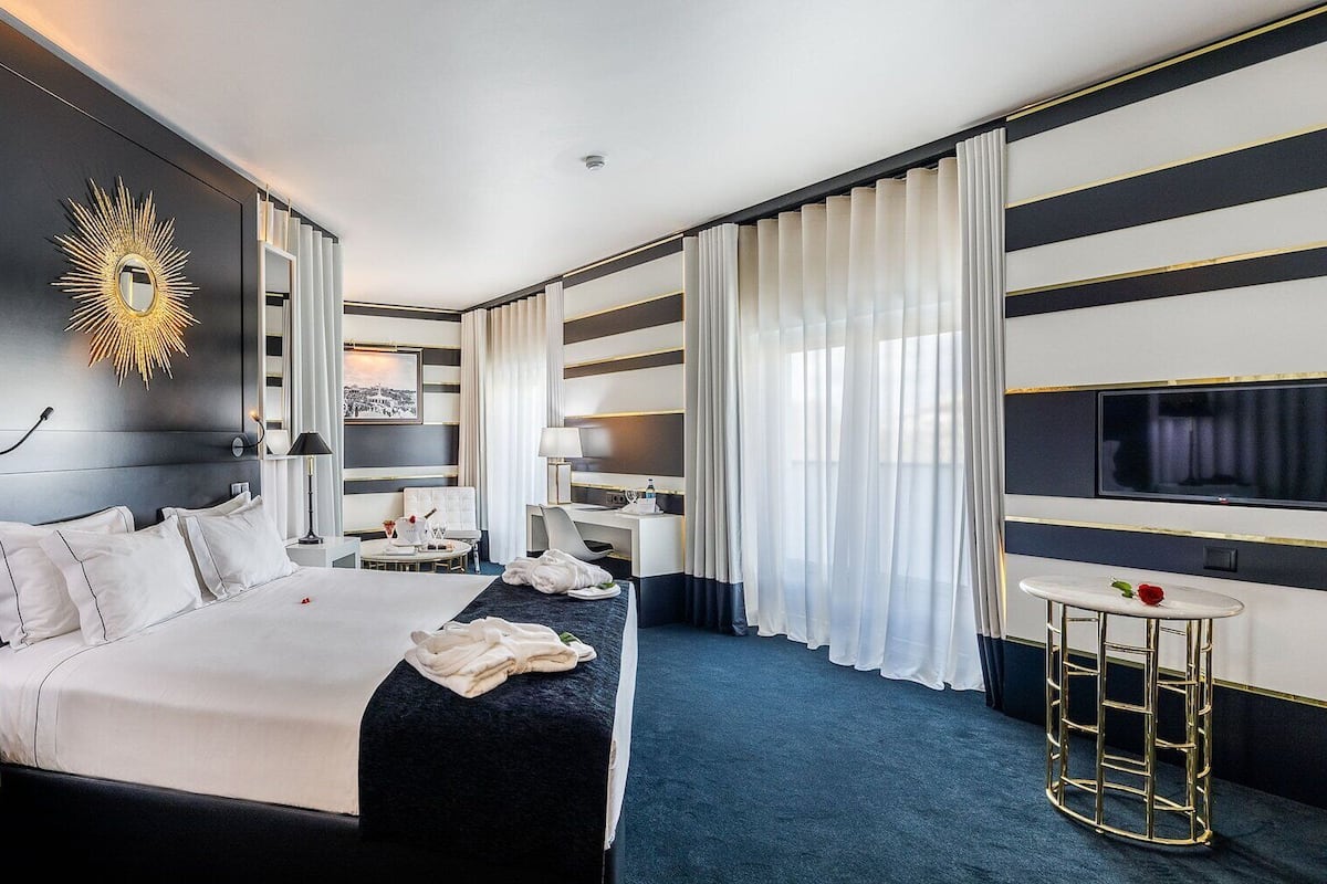 A modern hotel room with a large bed, bedside tables, a desk, a chair, a TV, and a decorative sunburst mirror.