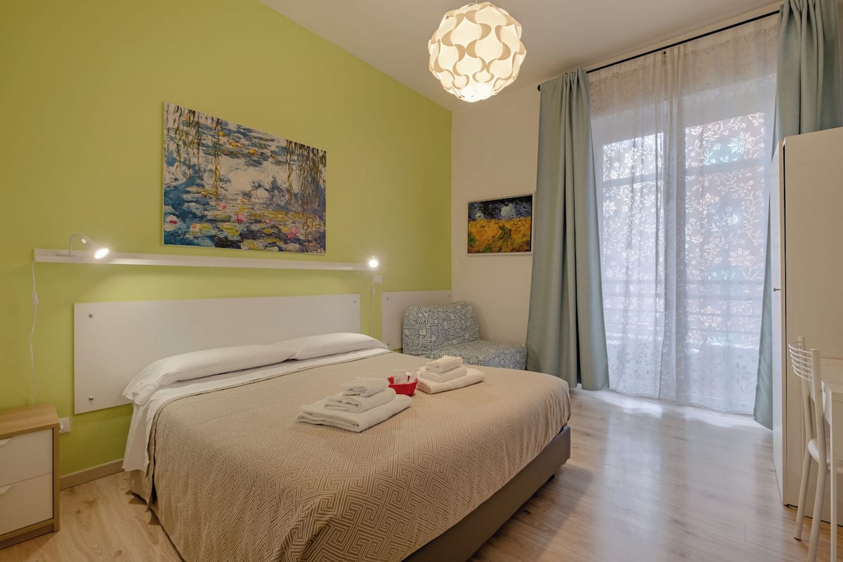 Ein Hotelzimmer mit Bett, einem Wandbild, einem Fenster mit Vorhängen und einer Deckenleuchte.